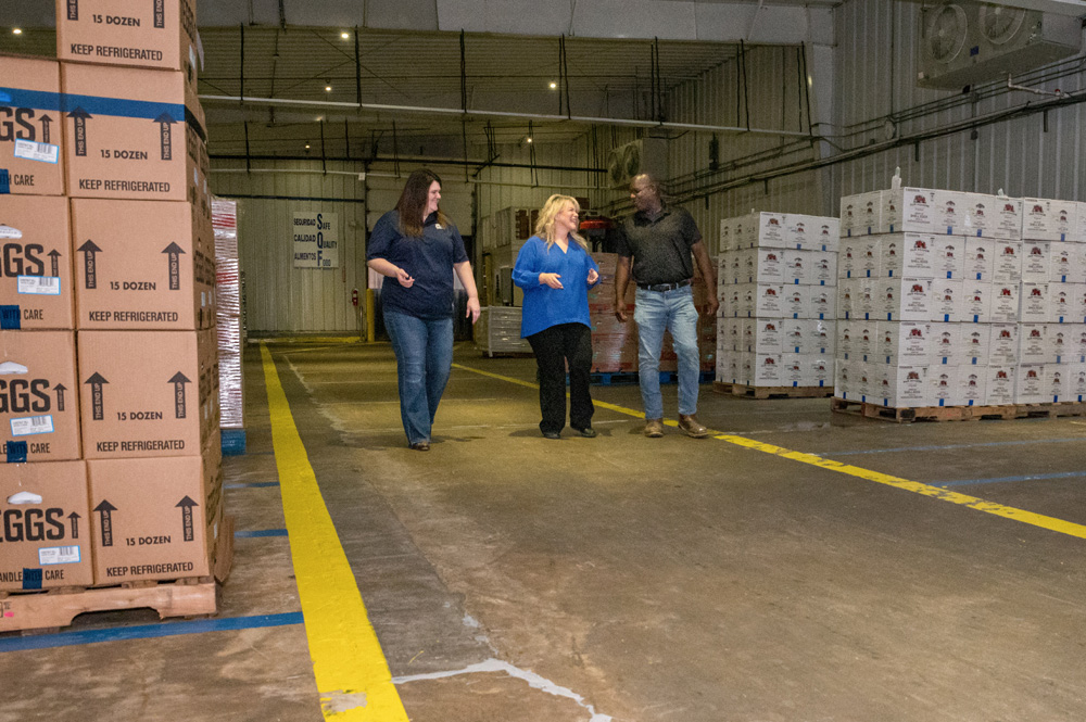 Three people walking through a loading dock full of crates of eggs. 