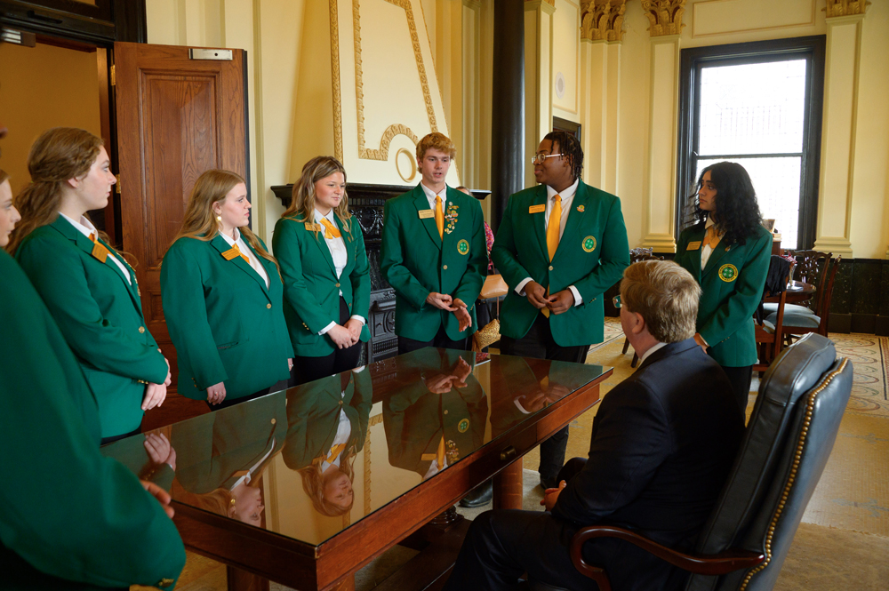 Six teens wearing green jackets talk with a seated person at a wooden desk.