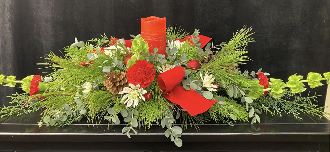 The completed design features multiple types of evergreens, red and white flowers, pine cones, a red ribbon, and a red candle in the center.