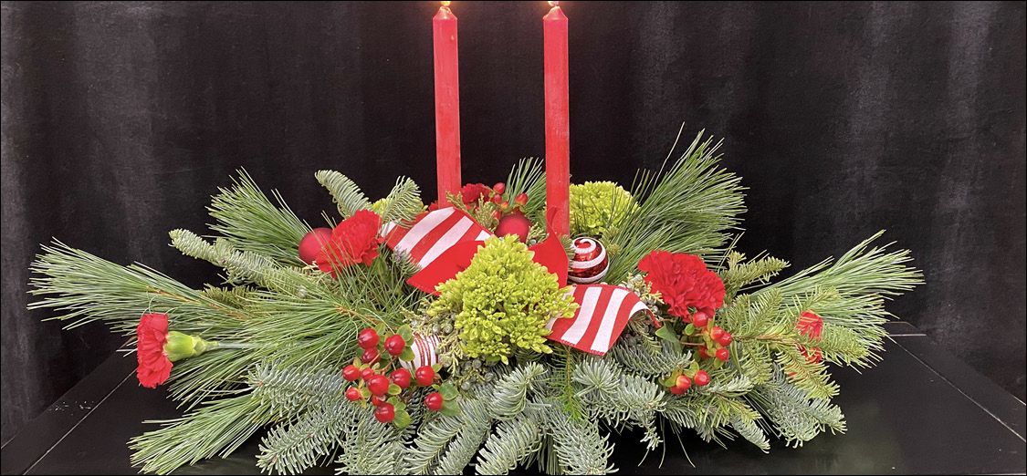 The completed design on a black table features different types of greenery, red berries, flowers, and ornaments, and two burning red candles.