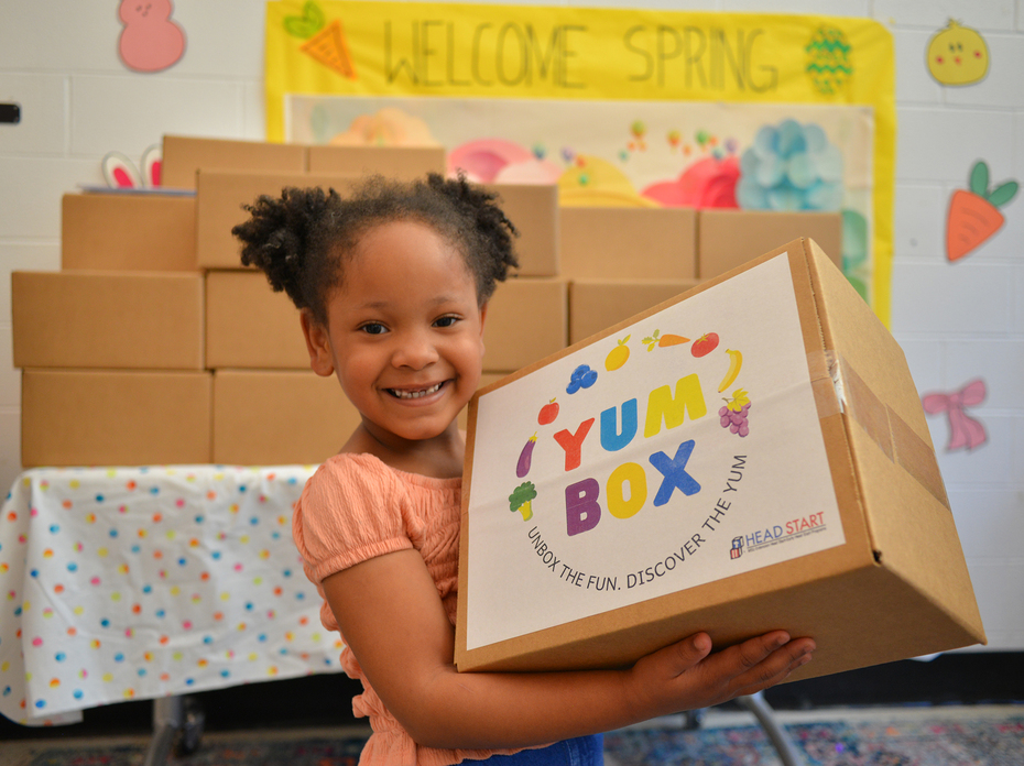 A young girl smiles while holding a colorful “Yum Box” package from Head Start, with stacked cardboard boxes and a “Welcome Spring” bulletin board visible behind her.