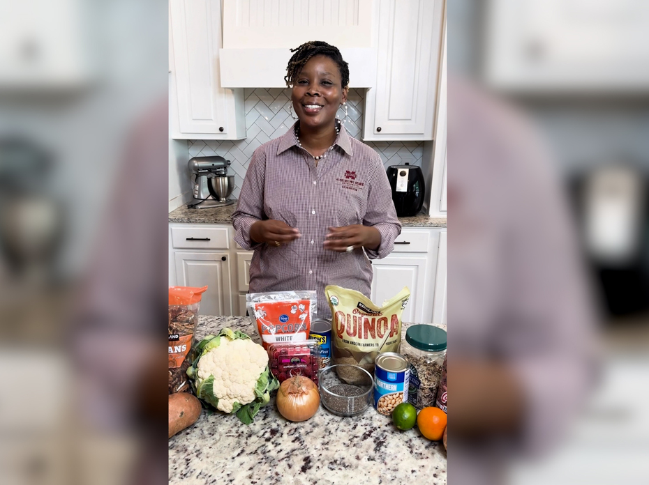 A woman stands behind a kitchen counter with several examples of fiber-rich foods displayed.