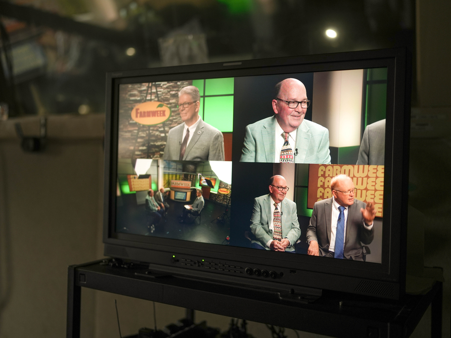Video monitor displays a talk show set with two men in suits being interviewed, shown from multiple camera angles, with a “Farmweek” sign in the studio background.