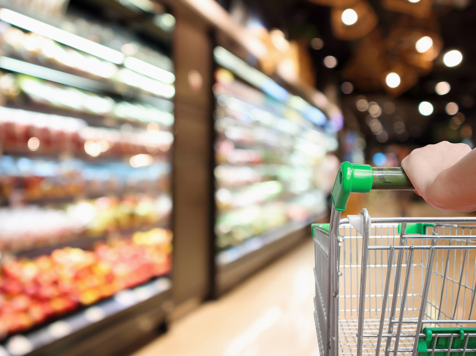 Woman’s hands hold a supermarket grocery cart with blurred fruits and vegetables in the background.