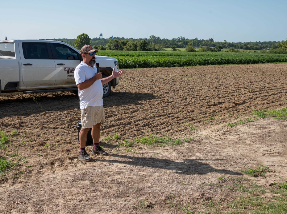 A speaker stands in front of research plots with a Mississippi State University Extension pickup truck in the background.