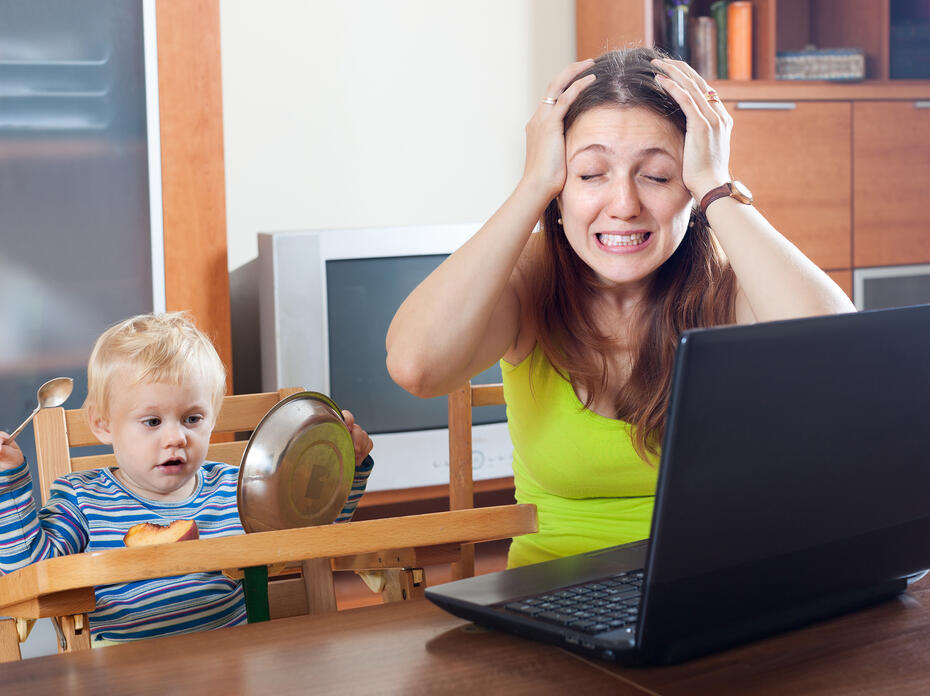 A young mother grabs her head in frustration while working from home on a laptop computer as her baby looks on. 
