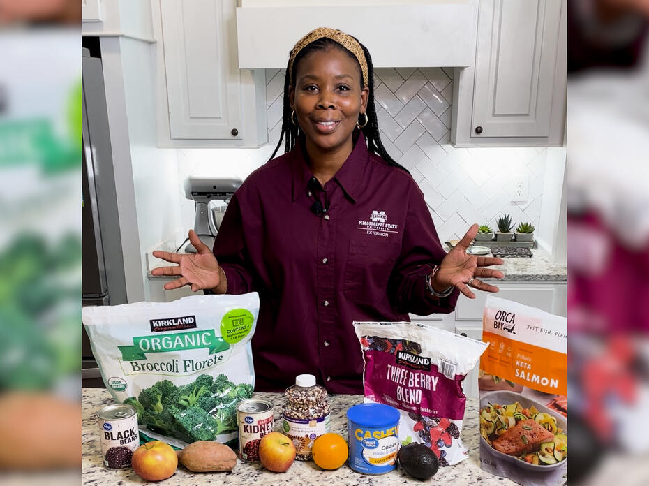 A woman stans in a kitchen with fruits, vegetables, fish, nuts and other foods on display.