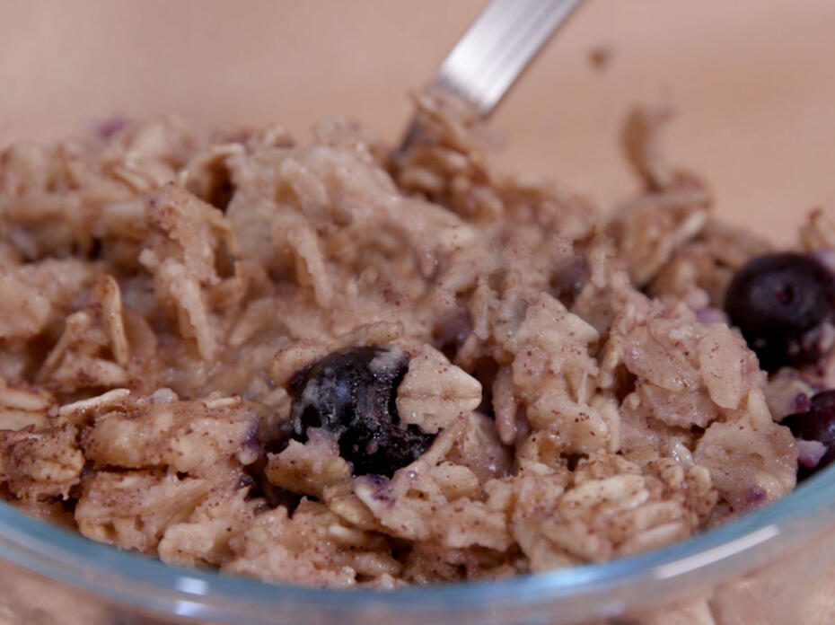 A small clear glass bowl holds baked oatmeal with blueberries and a stainless steel spoon.