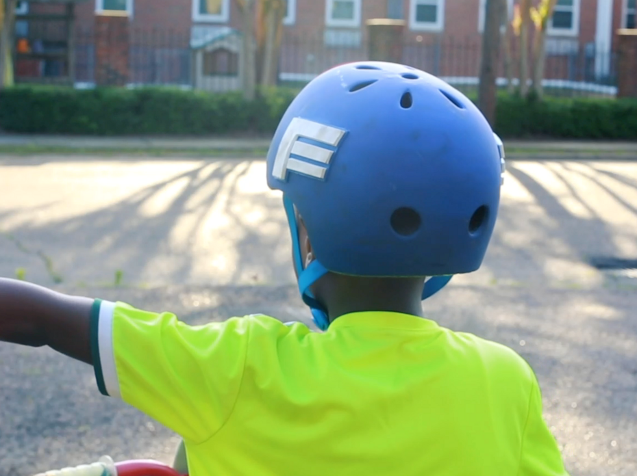 An African-American child wearing a blue helmet and neon yellow shirt extends his left hand to demonstrate a hand signal while riding a bicycle.