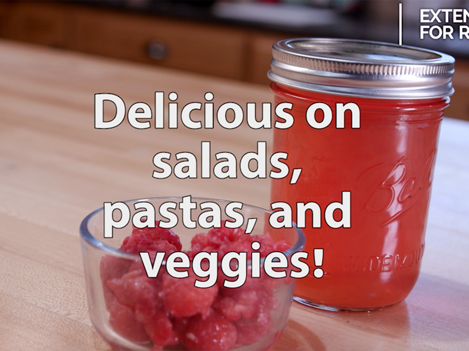A clear glass bowl of red raspberries sits on a butcher block table in front of a pint clear glass jar filled with homemade raspberry vinegar and topped with a metal lid and ring. Text overlay reads, "Delicious on salads, pastas, and veggies!"