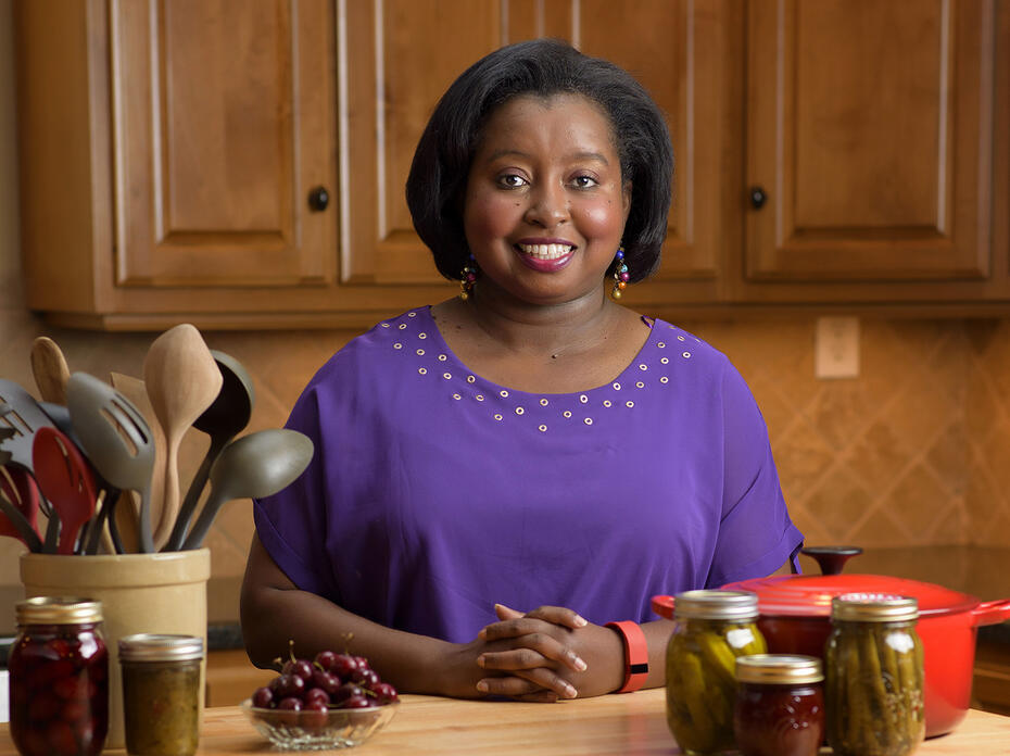 MSU Extension Agent Natasha Haynes, an African American woman with chin-length straight black hair stands behind a kitchen table displaying a crock of kitchen utensils, a red pot, and assorted home-canned and fresh fruits and vegetables. 