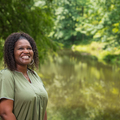 A person standing in front of a pond smiles at the sky.