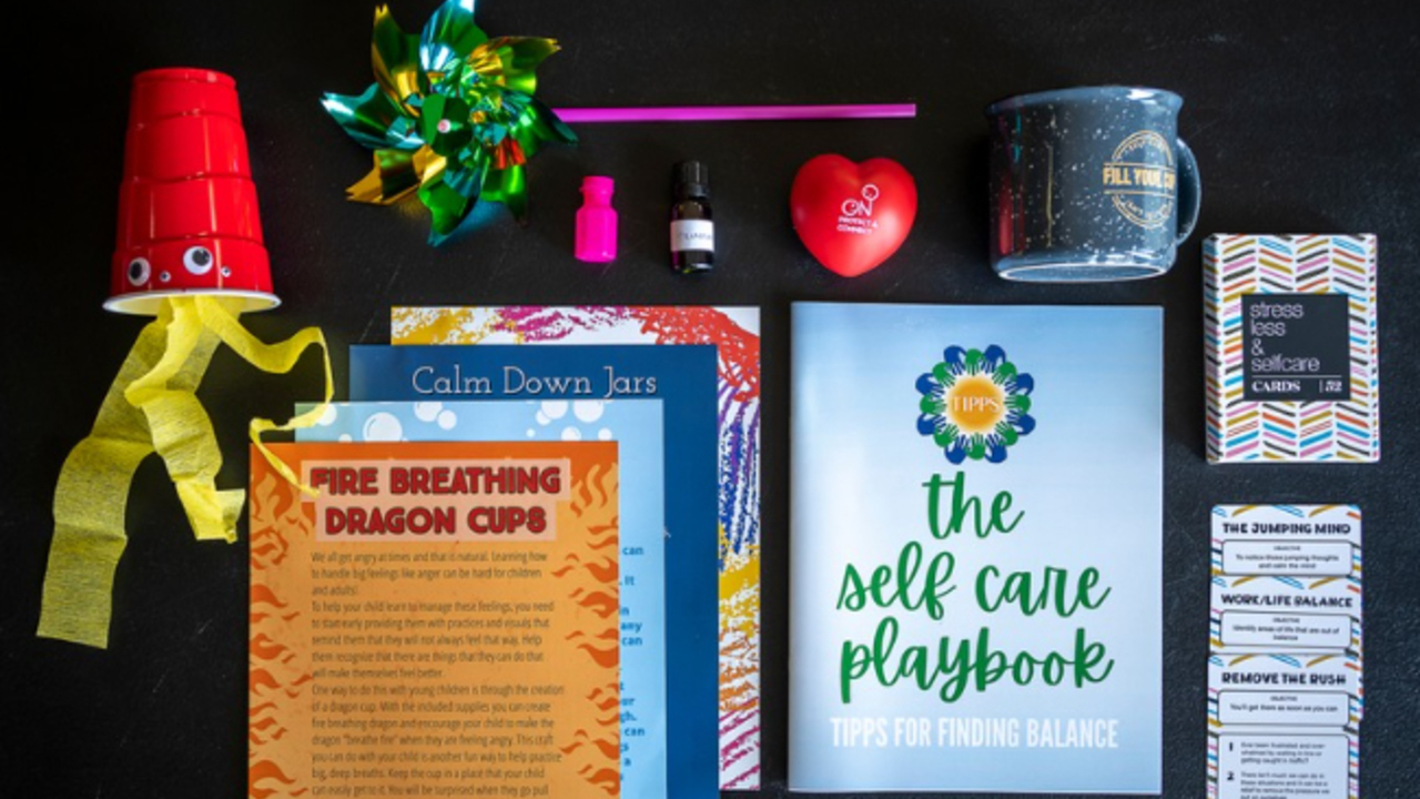 The contents of one toolkit, including a homemade fire-breathing dragon cup, various literature on parental self-care, strategies for emotion regulation and directions for crafts that support hand-eye coordination and fine motor skills, along with a coffee mug, essential oils, handheld pinwheel, a stress ball and bubbles. 