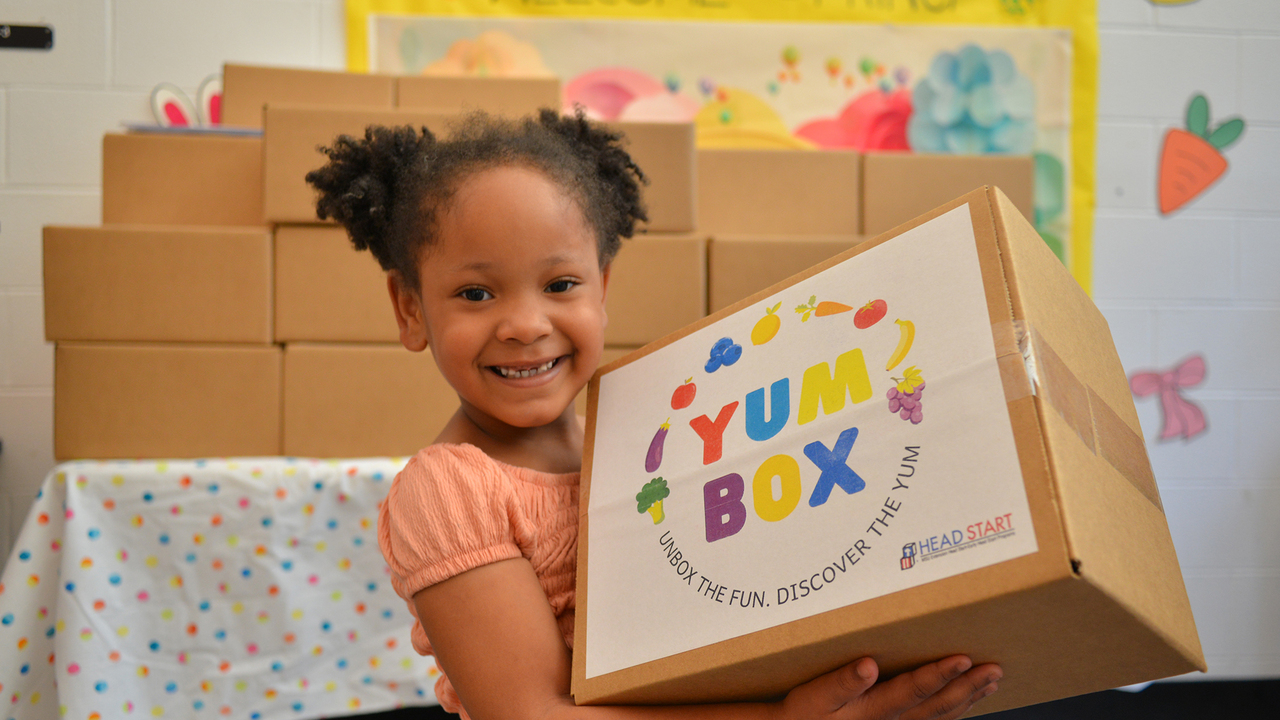 A young girl smiles while holding a colorful “Yum Box” package from Head Start, with stacked cardboard boxes and a “Welcome Spring” bulletin board visible behind her.