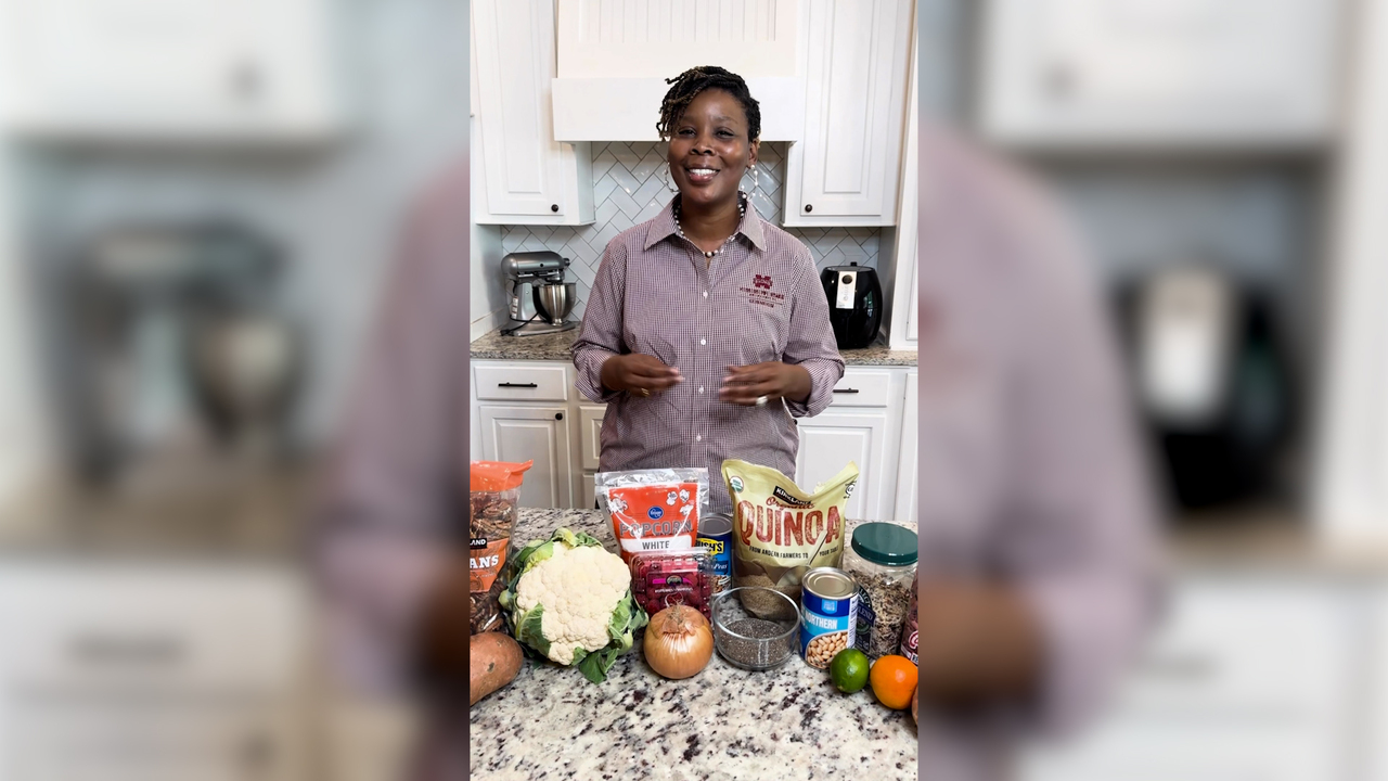 A woman stands behind a kitchen counter with several examples of fiber-rich foods displayed.