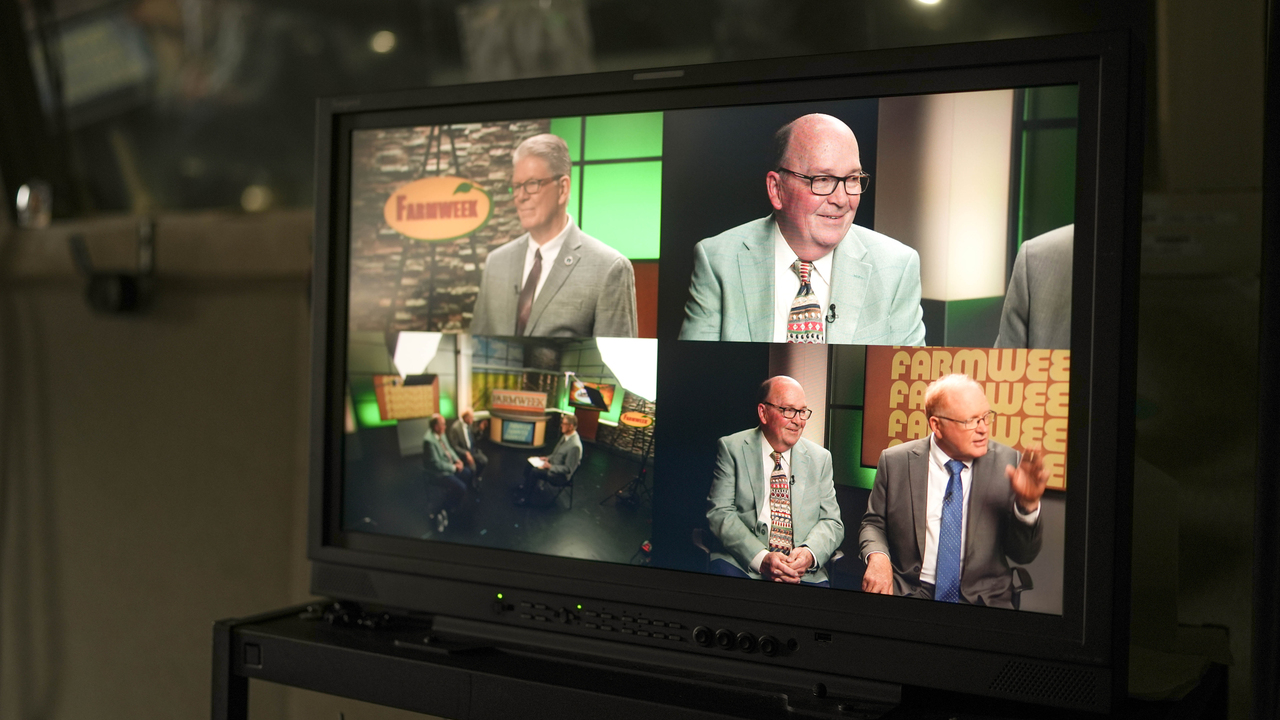 Video monitor displays a talk show set with two men in suits being interviewed, shown from multiple camera angles, with a “Farmweek” sign in the studio background.