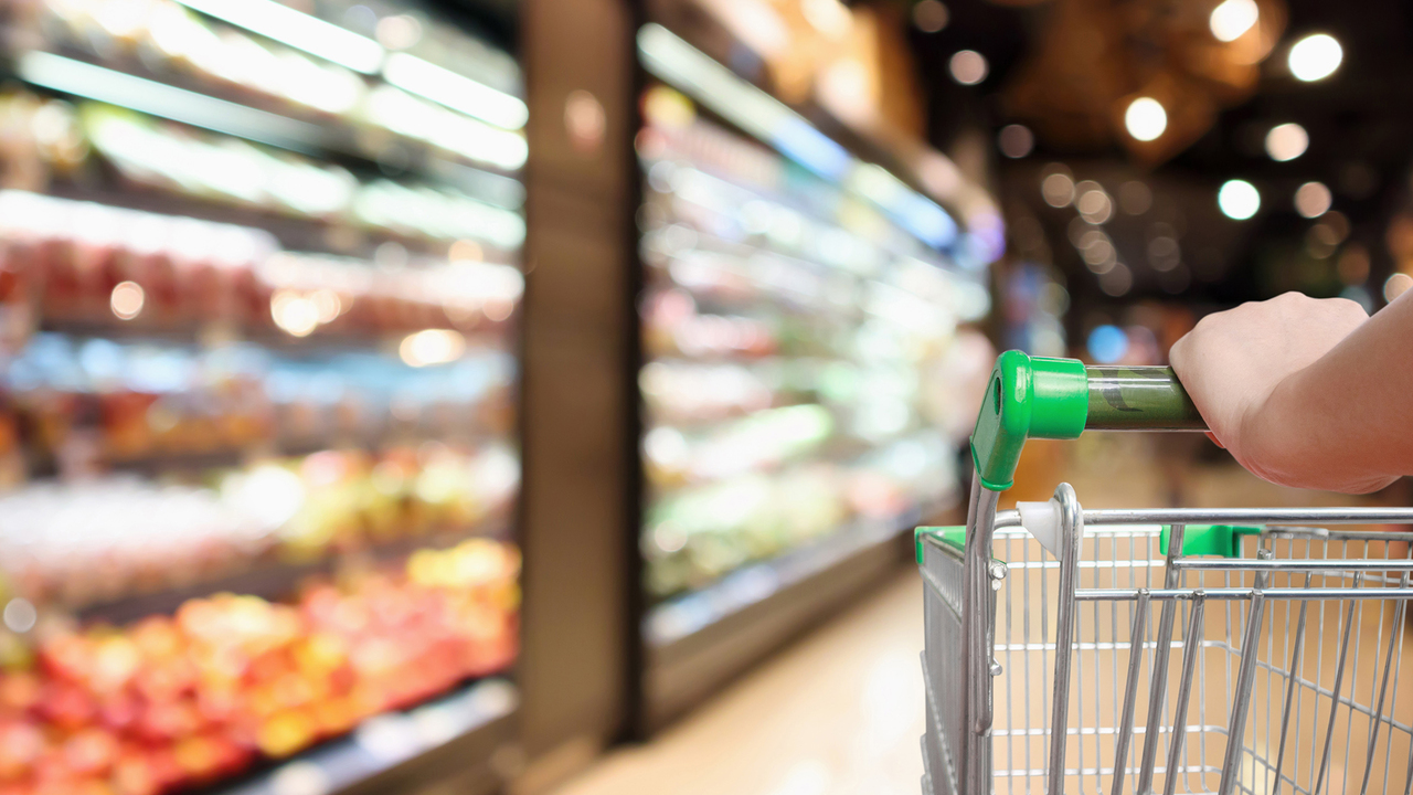 Woman’s hands hold a supermarket grocery cart with blurred fruits and vegetables in the background.