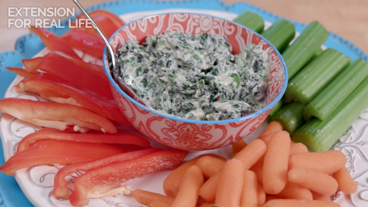 Spinach dip in a ceramic bowl centered on a plate surrounded by cut celery, baby carrots, and sliced red bell pepper.