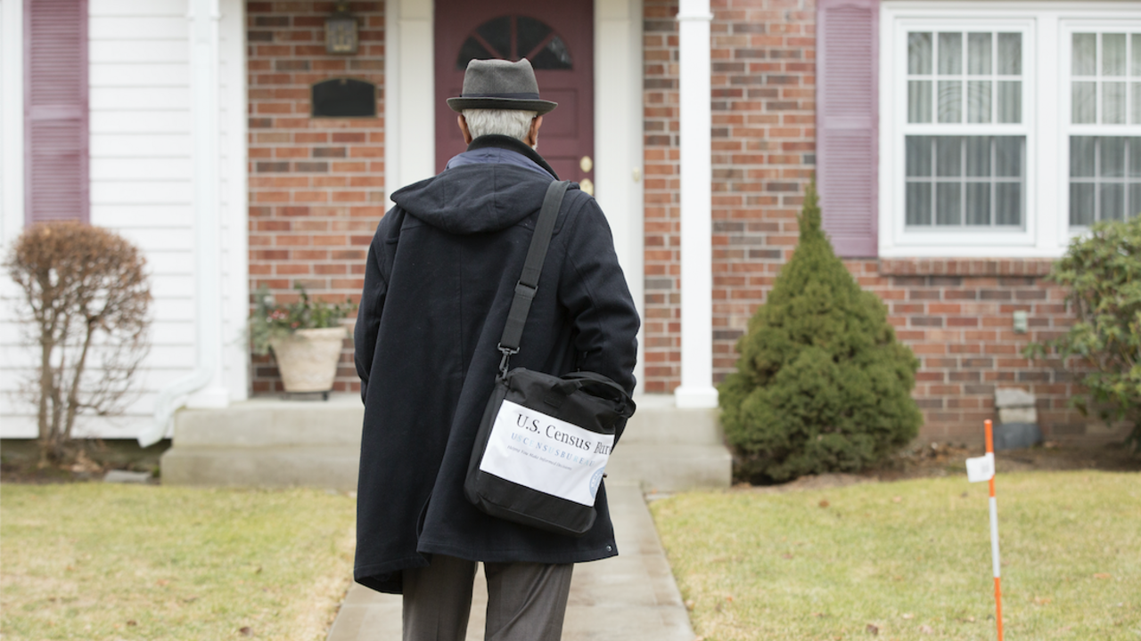 A census worker wearing a black hat and coat walking up to a house.