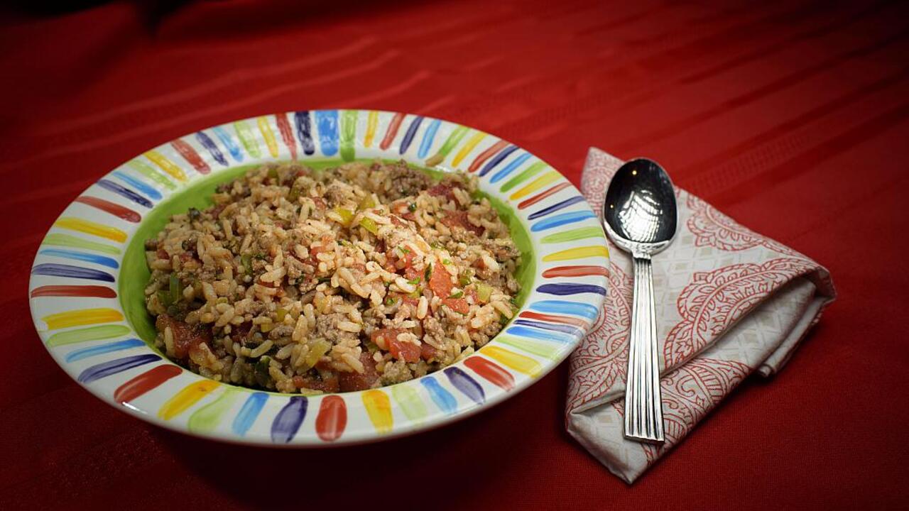 A bowl of Italian ground beef and rice in a rainbow-striped bowl sits on a red tablecloth with a red and white cloth napkin and a spoon.