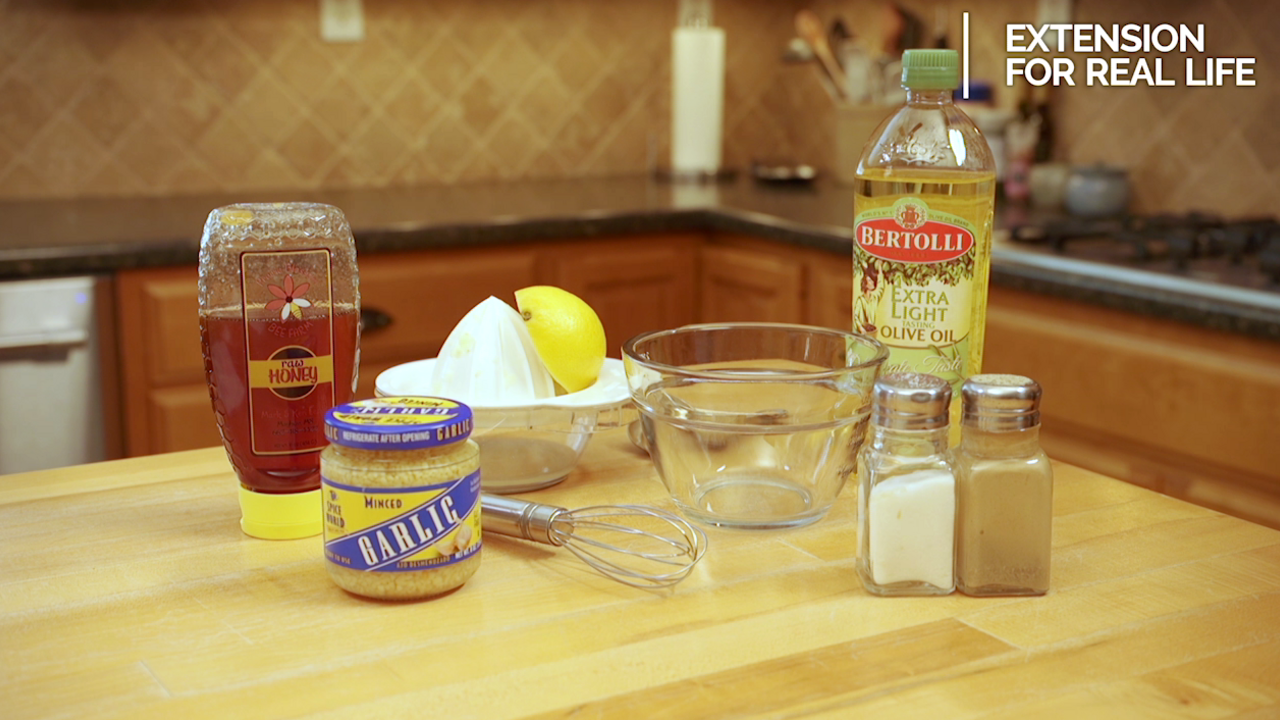 The ingredients and equipment to make honey lemon vinaigrette are displayed on a kitchen table, including honey, olive oil, half of a lemon, salt, pepper, minced garlic, a small glass bowl, a stainless steel whisk, and a citrus juicer. 