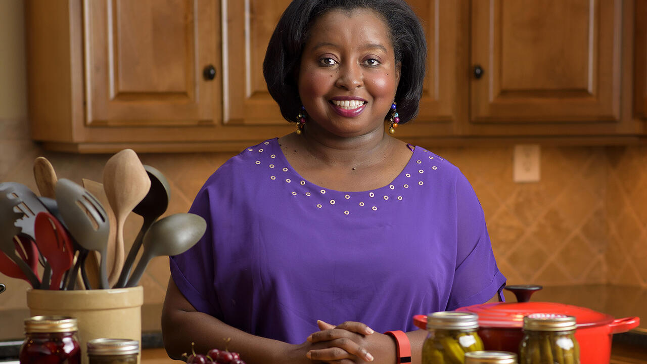 MSU Extension Agent Natasha Haynes, an African American woman with chin-length straight black hair stands behind a kitchen table displaying a crock of kitchen utensils, a red pot, and assorted home-canned and fresh fruits and vegetables. 