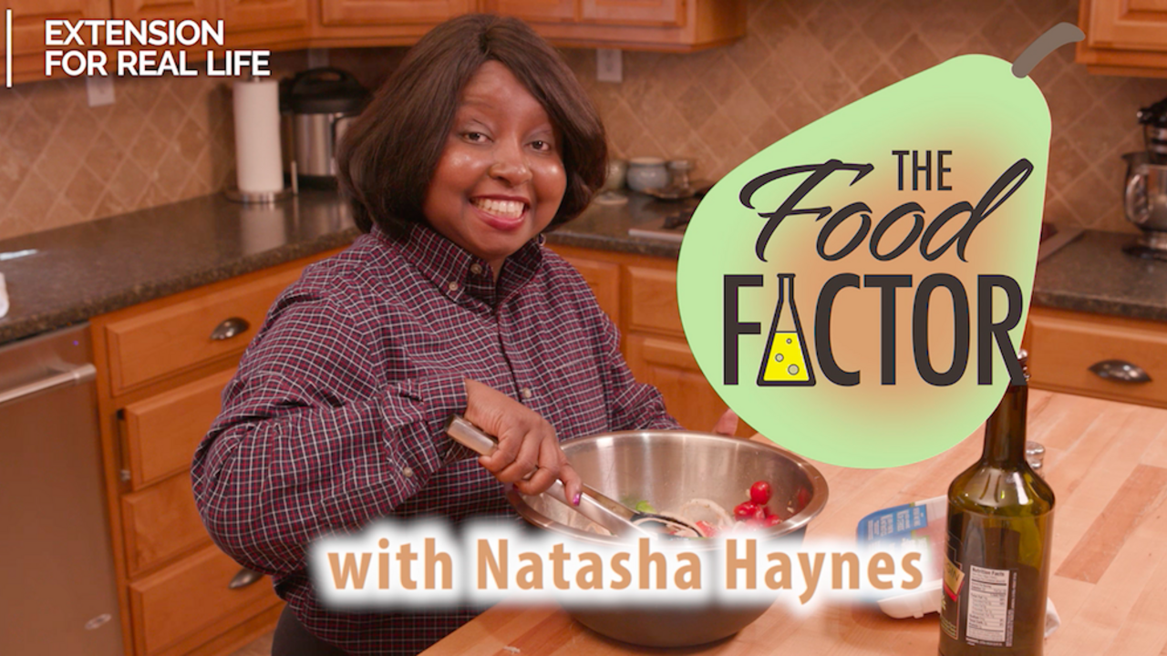 Natasha Haynes, an African American female wearing a plaid dress shirt stands in a kitchen and uses tongs to mix food ingredients in a stainless steel bowl. A tall bottle of olive oil is on the table. 
