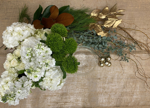 A glass bowl, gold salal, cluster of gold ornaments, white flowers, green florets, eucalyptus, and green foliage are laid out on burlap.