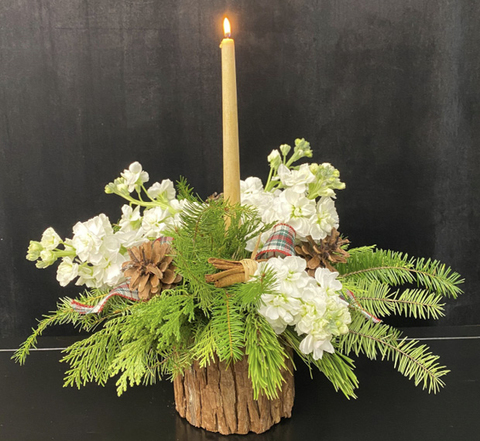 The completed design features two types of evergreen, white flowers, pine cones, and a single off-white candle burning in the center.