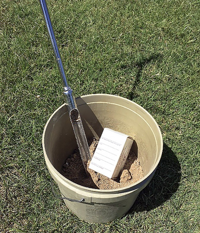 A plastic buckets half-filled with soil, with a soil probe and soil sample container on top.