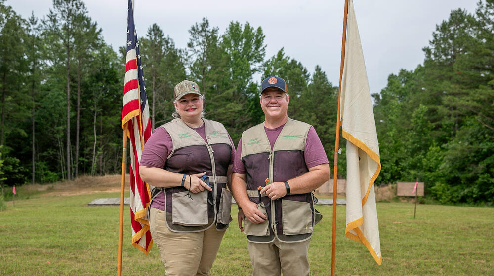 Two people, smiling and wearing 4-H Shooting Sports vests flanked by flags.